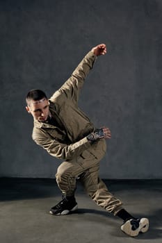 Good-looking fellow with tattooed body and face, earrings, beard. Dressed in khaki jumpsuit and black sneakers. He dancing against gray studio background. Dancehall, hip-hop. Full length, copy space