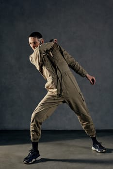 Young stately male with tattooed body and face, earrings, beard. Dressed in khaki jumpsuit and black sneakers. He is dancing against gray studio background. Dancehall, hip-hop. Full length, copy space