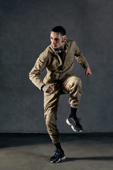 Young stately performer with tattooed body and face, earrings, beard. Dressed in khaki overalls and black sneakers. He is dancing against gray background. Dancehall, hip-hop. Full length, copy space