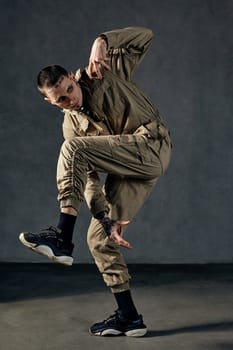 Young strong guy with tattooed body and face, earrings, beard. Dressed in khaki overalls and black sneakers. He is dancing against gray studio background. Dancehall, hip-hop. Full length, copy space