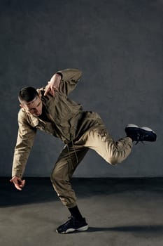 Young strong fellow with tattooed body and face, earrings, beard. Dressed in khaki jumpsuit and black sneakers. He dancing against gray studio background. Dancehall, hip-hop. Full length, copy space