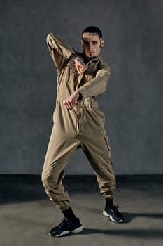 Young active man with tattooed body and face, earrings, beard. Dressed in khaki overalls and black sneakers. He is dancing against gray studio background. Dancehall, hip-hop. Full length, copy space