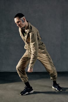 Young active fellow with tattooed body and face, earrings, beard. Dressed in khaki jumpsuit and black sneakers. He dancing against gray studio background. Dancehall, hip-hop. Full length, copy space