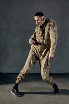 Young active performer with tattooed body and face, earrings, beard. Dressed in khaki overalls and black sneakers. He is dancing against gray background. Dancehall, hip-hop. Full length, copy space