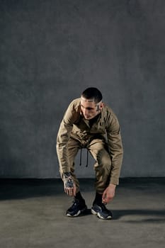 Young flexible man with tattooed body and face, earrings, beard. Dressed in khaki overalls and black sneakers. He is dancing against gray studio background. Dancehall, hip-hop. Full length, copy space