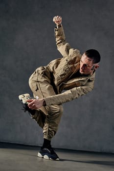 Young attractive male with tattooed body and face, earrings, beard. Dressed in khaki jumpsuit and black sneakers. He dancing against gray studio background. Dancehall, hip-hop. Full length, copy space