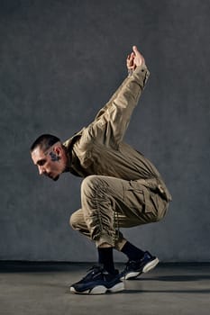 Young attractive fellow with tattooed body and face, earrings, beard. Dressed in khaki jumpsuit and black sneakers. He dancing against gray background. Dancehall, hip-hop. Full length, copy space