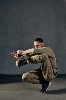 Young unusual fellow with tattooed body and face, earrings, beard. Dressed in khaki jumpsuit and black sneakers. He dancing against gray studio background. Dancehall, hip-hop. Full length, copy space