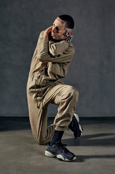 Young unusual performer with tattooed body and face, earrings, beard. Dressed in khaki overalls and black sneakers. He is dancing against gray background. Dancehall, hip-hop. Full length, copy space