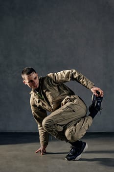 Extraordinary male with tattooed body and face, earrings, beard. Dressed in khaki jumpsuit and black sneakers. He is dancing against gray studio background. Dancehall, hip-hop. Full length, copy space