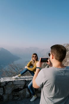 Man takes a picture with a smartphone of woman sitting on a fence over the Bay of Kotor. Montenegro. Back view. High quality photo