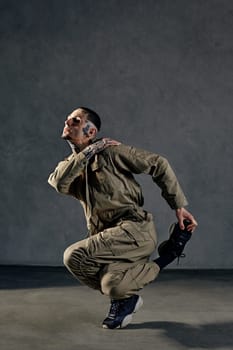 Extraordinary guy with tattooed body and face, earrings, beard. Dressed in khaki overalls and black sneakers. He is dancing against gray studio background. Dancehall, hip-hop. Full length, copy space
