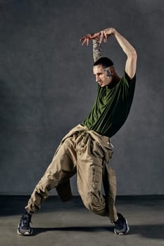 Young stylish fellow with tattooed body and face, earrings, beard. Dressed in khaki t-shirt and jumpsuit, black sneakers. Dancing against gray background. Dancehall, hip-hop. Full length, copy space