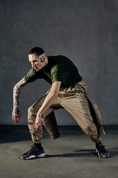 Young graceful man with tattooed body and face, earrings, beard. Dressed in green t-shirt and khaki overalls, black sneakers. Dancing on gray background. Dancehall, hip-hop. Full length, copy space