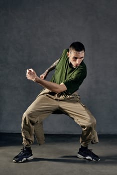 Young graceful male with tattooed body and face, earrings, beard. Dressed in khaki t-shirt and jumpsuit, black sneakers. Dancing against gray background. Dancehall, hip-hop. Full length, copy space