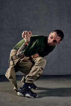 Young graceful fellow with tattooed body and face, earrings, beard. Dressed in khaki t-shirt and jumpsuit, black sneakers. Dancing against gray background. Dancehall, hip-hop. Full length, copy space