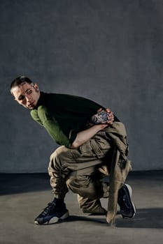 Young graceful performer with tattooed body and face, earrings, beard. Dressed in khaki t-shirt and overalls, black sneakers. Dancing on gray background. Dancehall, hip-hop. Full length, copy space