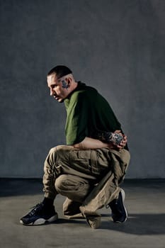 Young flexible man with tattooed body and face, earrings, beard. Dressed in green t-shirt and khaki overalls, black sneakers. Dancing on gray background. Dancehall, hip-hop. Full length, copy space