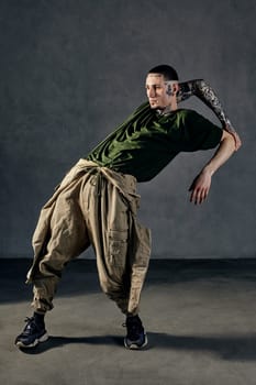 Young flexible guy with tattooed body and face, earrings, beard. Dressed in green t-shirt and khaki overalls, black sneakers. Dancing on gray background. Dancehall, hip-hop. Full length, copy space
