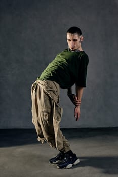 Young athletic male with tattooed body and face, earrings, beard. Dressed in khaki t-shirt and jumpsuit, black sneakers. Dancing against gray background. Dancehall, hip-hop. Full length, copy space