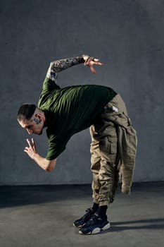 Young athletic guy with tattooed body and face, earrings, beard. Dressed in green t-shirt and khaki overalls, black sneakers. Dancing on gray background. Dancehall, hip-hop. Full length, copy space