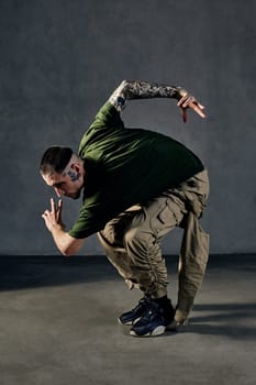 Young athletic fellow with tattooed body and face, earrings, beard. Dressed in khaki t-shirt and jumpsuit, black sneakers. Dancing against gray background. Dancehall, hip-hop. Full length, copy space