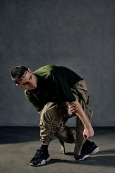 Young athletic performer with tattooed body and face, earrings, beard. Dressed in khaki t-shirt and overalls, black sneakers. Dancing on gray background. Dancehall, hip-hop. Full length, copy space