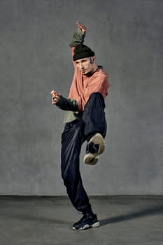Young strong performer with tattooed body and face, earrings, beard. Dressed in hat, casual clothes and black sneakers. Dancing on gray background. Dancehall, hip-hop. Full length, copy space