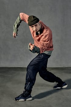 Young graceful guy with tattooed body and face, earrings, beard. Dressed in hat, casual clothes and black sneakers. Dancing on gray background. Dancehall, hip-hop. Full length, copy space