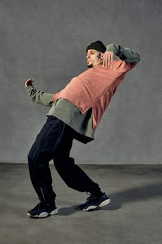 Graceful fellow with tattooed body and face, earrings, beard. Dressed in hat, colorful jumper, black pants and sneakers. Dancing on gray background. Dancehall, hip-hop. Full length, copy space