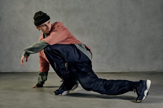 Modern fellow with tattooed body and face, earrings, beard. Dressed in hat, colorful jumper, black pants and sneakers. Dancing on gray background. Dancehall, hip-hop. Full length, copy space