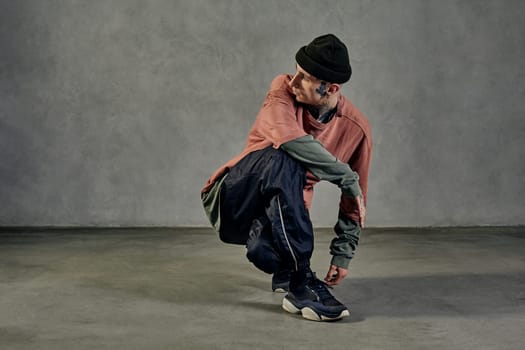 Young modern performer with tattooed body and face, earrings, beard. Dressed in hat, casual clothes and black sneakers. Dancing on gray background. Dancehall, hip-hop. Full length, copy space