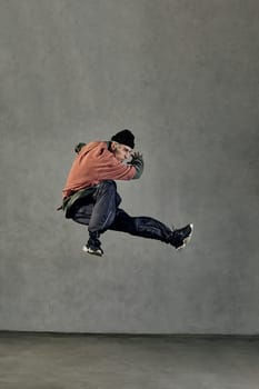 Young athletic performer with tattooed face, earrings, beard. Dressed in hat, casual clothes and black sneakers. Jumping while dancing on gray background. Dancehall, hip-hop. Full length, copy space