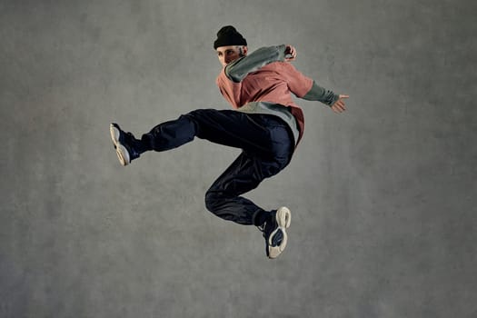 Athletic fellow with tattooed face, earrings, beard. Dressed in hat, colorful jumper, black pants and sneakers. Jumping while dancing on gray background. Dancehall, hip-hop. Full length, copy space