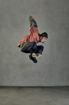 Young stately man with tattooed body and face, earrings, beard. Dressed in hat, casual clothes and black sneakers. Jumping while dancing on gray background. Dancehall, hip-hop. Full length, copy space