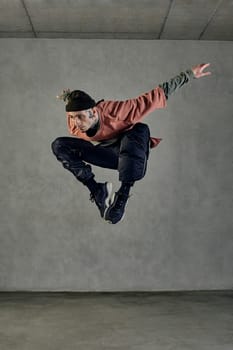 Young stately guy with tattooed body and face, earrings, beard. Dressed in hat, casual clothes and black sneakers. Jumping while dancing on gray background. Dancehall, hip-hop. Full length, copy space