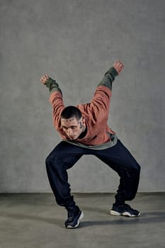 Young flexible man with tattooed body and face, earrings, beard. Dressed in casual clothes and black sneakers. Dancing on gray background. Dancehall, hip-hop. Full length, copy space