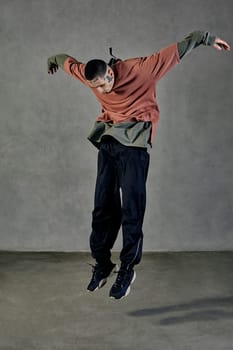 Young flexible guy with tattooed body and face, earrings, beard. Dressed in casual clothes and black sneakers. Jumping while dancing on gray background. Dancehall, hip-hop. Full length, copy space