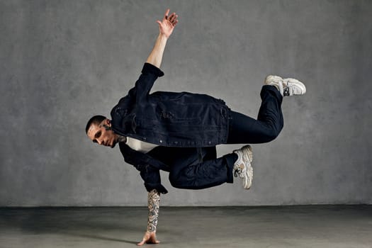 Active male with tattooed body and face, earrings, beard. Dressed in white t-shirt and sneakers, black denim shirt, pants. Dancing on gray background. Dancehall, hip-hop. Full length, copy space