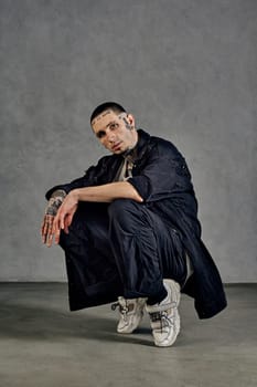 Active fellow with tattooed body and face, earrings, beard. Dressed in white t-shirt and sneakers, black denim shirt, pants. Squatting on socks against gray background. Dancehall, hip-hop. Close up