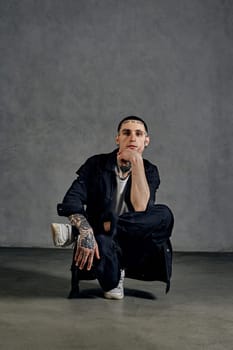 Stylish man with tattooed body, earrings, beard. Dressed in white t-shirt and sneakers, black denim shirt and pants. Performing tricks on gray background. Dancehall, hip-hop. Close up, copy space