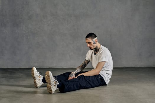 Athletic guy with tattooed body and face, earrings, beard. Dressed in white t-shirt and sneakers, black sports trousers. Sitting on floor against gray background. Dancehall, hip-hop. Close up