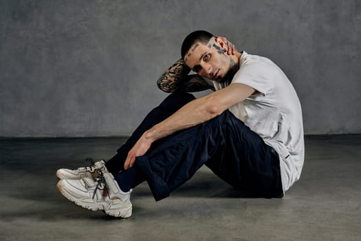 Performer with tattooed body and face, earrings, beard. Dressed in white t-shirt and sneakers, black sports trousers. Sitting on floor against gray background. Dancehall, hip-hop. Close up, side view