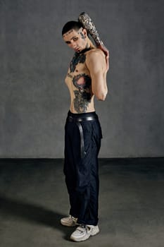 Strong fellow with tattooed body and face, naked torso, beard. Dressed in black pants and white sneakers. Stretching while posing sideways against gray background. Dancehall, hip-hop. Full length