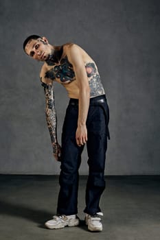 Flexible man with tattooed body and face, naked torso, beard. Dressed in black pants and white sneakers. He is dancing against gray studio background. Dancehall, hip-hop. Full length, copy space
