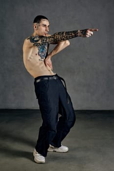 Flexible fellow with tattooed body and face, naked torso, beard. Dressed in black pants and white sneakers. He is dancing on gray studio background. Dancehall, hip-hop. Full length, copy space