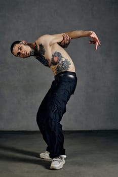 Athletic male with tattooed body and face, naked torso, beard. Dressed in black pants and white sneakers. He is dancing on gray studio background. Dancehall, hip-hop. Full length, copy space