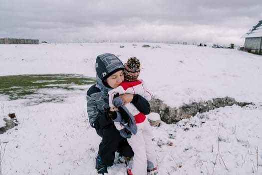 Mom hugs a little girl squatting on a snowy pasture near a farm. High quality photo