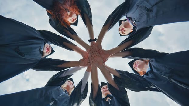 Team of college or university students celebrating graduation. Group of happy successful graduates in academic hats and robes standing in circle and putting their hands together