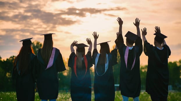 Silhouettes of graduates in black robes waving their arms against the evening sunset